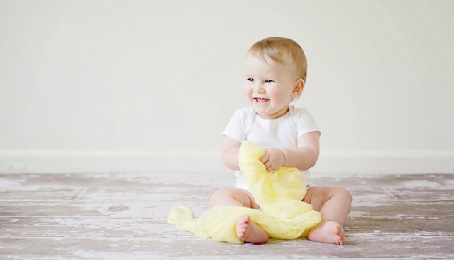 Smaidošs mazulis sēž uz grīdas, turpt rokā dzeltenu lakatu